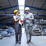 Manager supervisor and industrial worker in uniform walking in large metal factory hall and talking about increasing production.